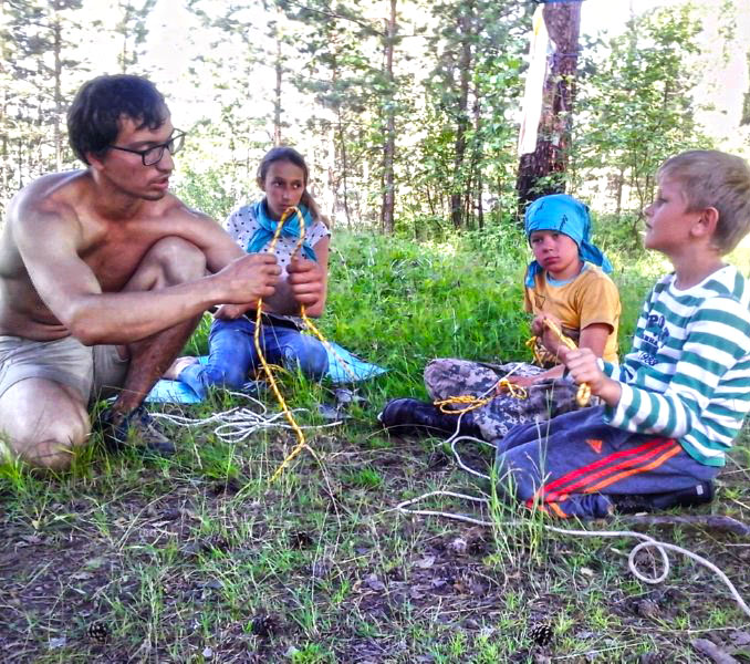 Взрослый в очках обучает детей вязанию морских узлов на траве в лесу в рамках программы "Сам Себе Робинзон" летом 2017 года.
